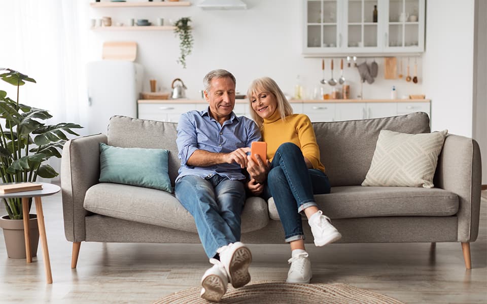 Happy Mature Husband And Wife Using Smartphone Sitting At Home