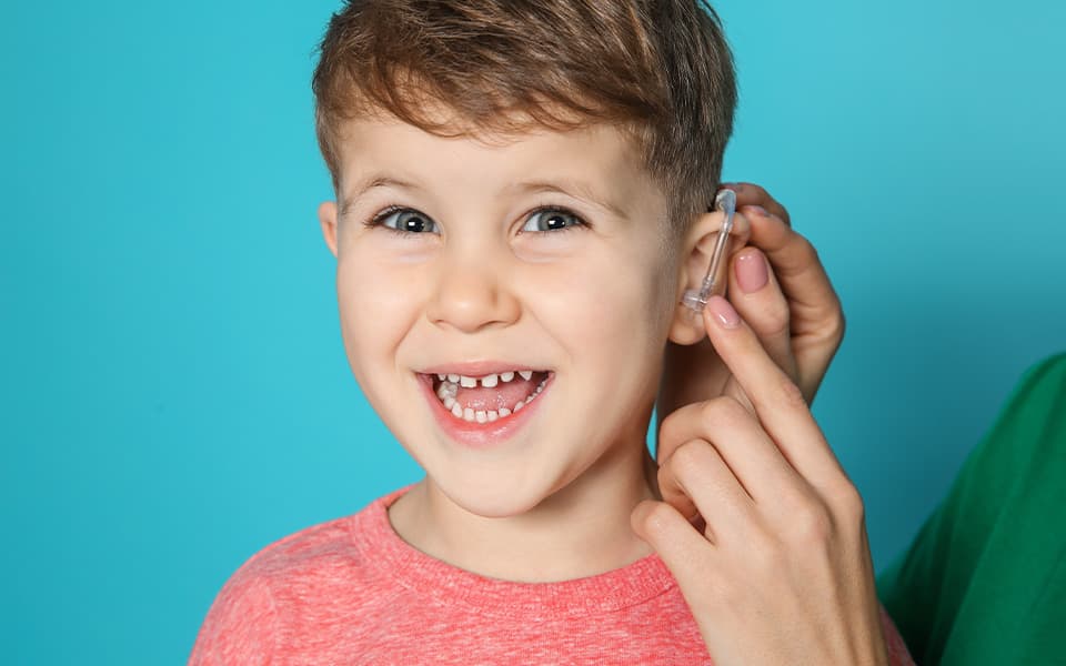 Young woman adjusting little son's hearing aid on color background