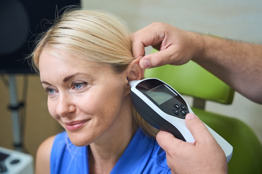Image of a woman with blond hair sitting in a chair while a doctor tugs her ear upward and inserts a tympanometer device