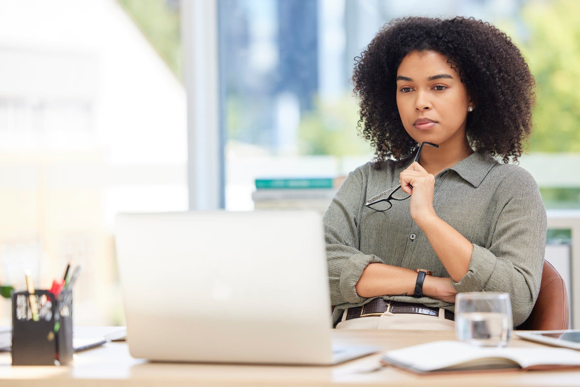 A professional woman with curly hair sits at an office desk, holding her glasses and looking thoughtfully at her laptop