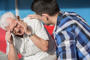 Senior man holding hands to head as if experiencing balance issues while younger man comforts him by placing a hand on the older man's shoulder