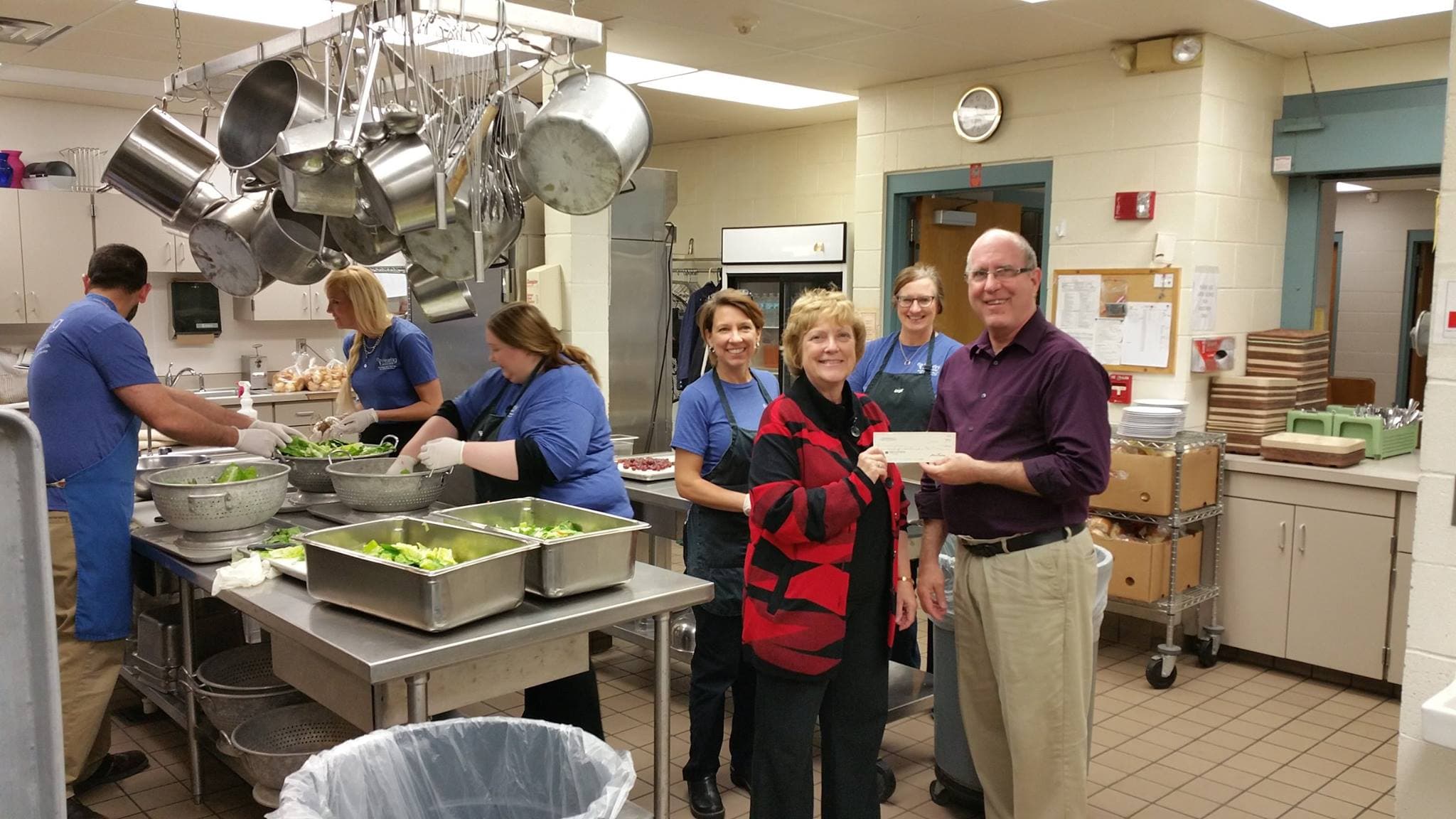 Man and woman holding a check in a community kitchen with worker's in the background preparing a salad